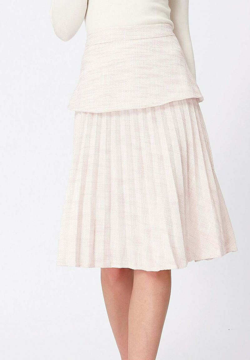 Jupe plissée à longueur genou en tissu texturé de couleur rose doux, portée avec un haut à manches longues crème sur fond blanc.
