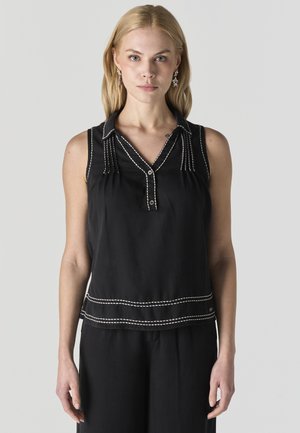 Mujer rubia con blusa negra sin mangas con costuras blancas, cuello en V y pantalones negros, posando frente a un fondo liso de color gris claro.