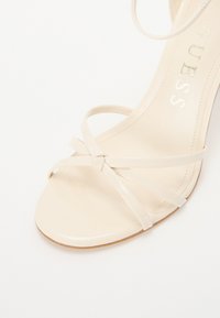 Beige high-heeled sandal with glossy patent finish, featuring crisscross straps and a smooth toe area. Brand name embossed on the insole.