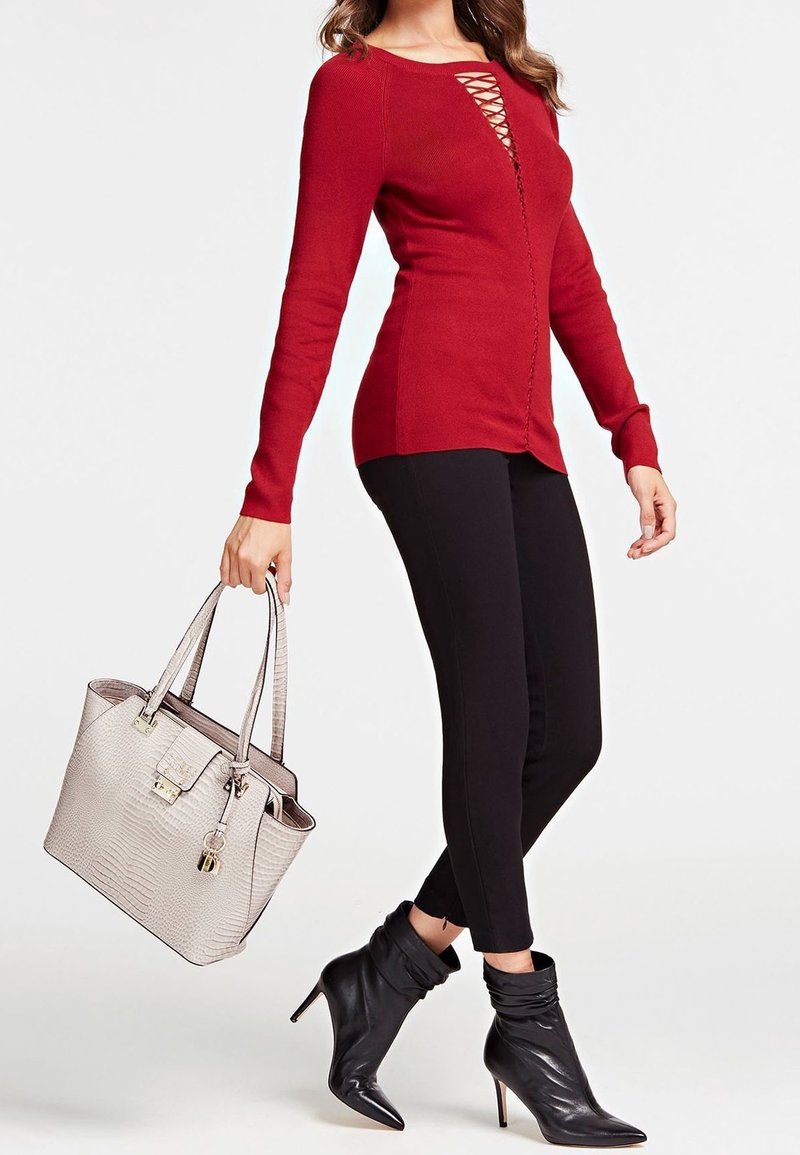 Femme portant un haut rouge à manches longues avec un détail en dentelle sur le devant, un pantalon noir slim, des bottines noires, tenant un sac à main beige texturé.