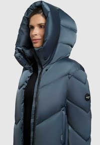 Giacca imbottita con cappuccio di colore blu scuro, caratterizzata da un design trapuntato, con grandi tasche e una patch rotonda con logo sulla manica sinistra.