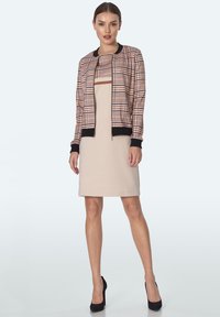 Kurtka bomber w kratę z czarnymi akcentami na dopasowanej beżowej sukience. Teksturowany materiał i zamek błyskawiczny; wysokie obcasy dopełniają stylizację.
