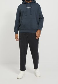 Granatowa bluza z kapturem z kieszenią z przodu i białym logo, zestawiona z czarnymi, zwężanymi spodniami i białymi trampkami, na prostym tle.