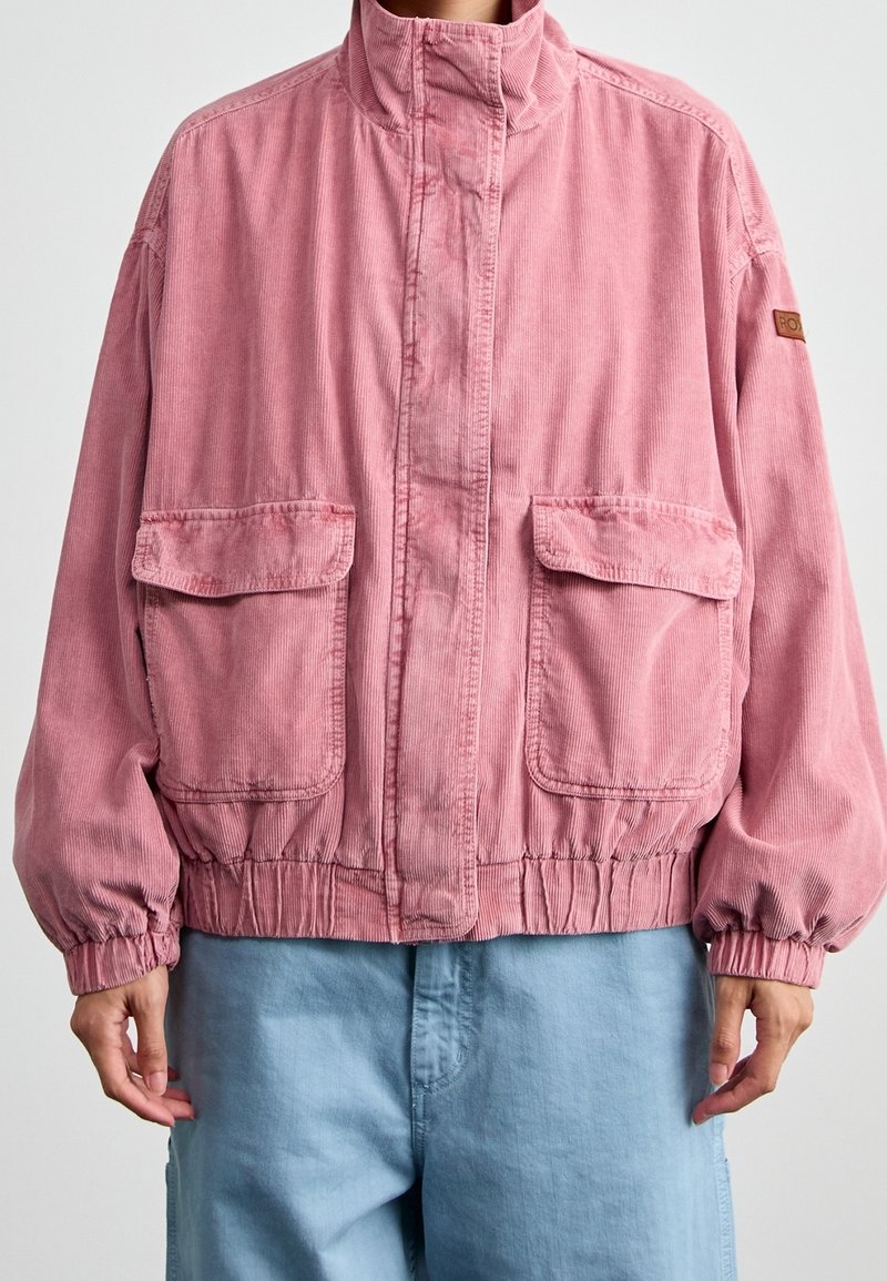 Personne portant une veste en velours côtelé rose avec de grandes poches avant à rabat et des poignets élastiques, associée à un pantalon bleu clair.