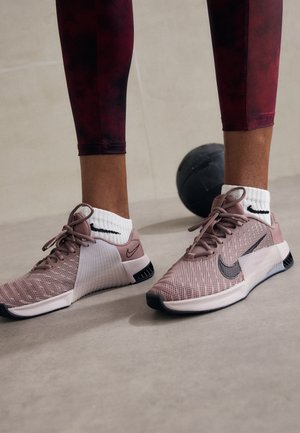 Osoba nosząca fioletowe buty sportowe Nike z białymi skarpetkami Nike i bordowymi legginsami stoi obok czarnej piłki lekarskiej na jasnej podłodze.