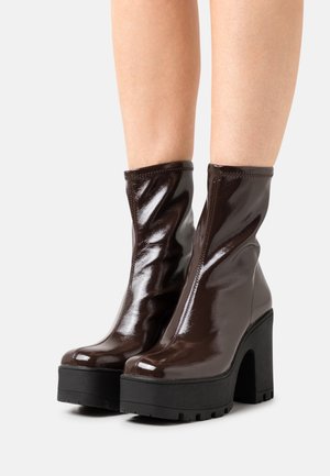 Jambes portant des bottines à plateforme marron foncé brillantes, avec des talons épais noirs et des semelles crantées, sur fond blanc.