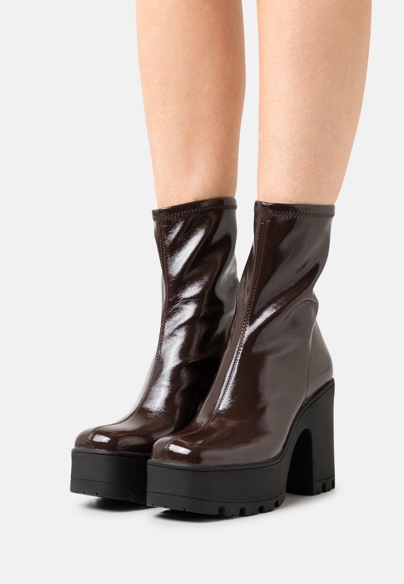 Jambes portant des bottines à plateforme marron foncé brillantes, avec des talons épais noirs et des semelles crantées, sur fond blanc.