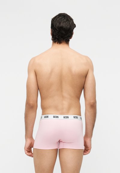 Άνδρας με κοντά σκούρα μαλλιά, όρθιος με την πλάτη προς τον θεατή, φορώντας ανοιχτό ροζ κοντά αντρικά εσώρουχα με λευκή μέση που φέρει το λογότυπο "GCDS".