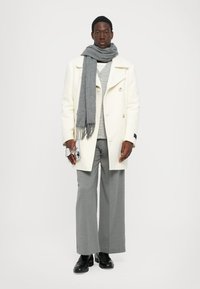 Cappotto bianco doppiopetto, maglione grigio a fantasia, pantaloni grigi a gamba larga, sciarpa grigia, scarpe nere, con in mano una piccola macchina fotografica.