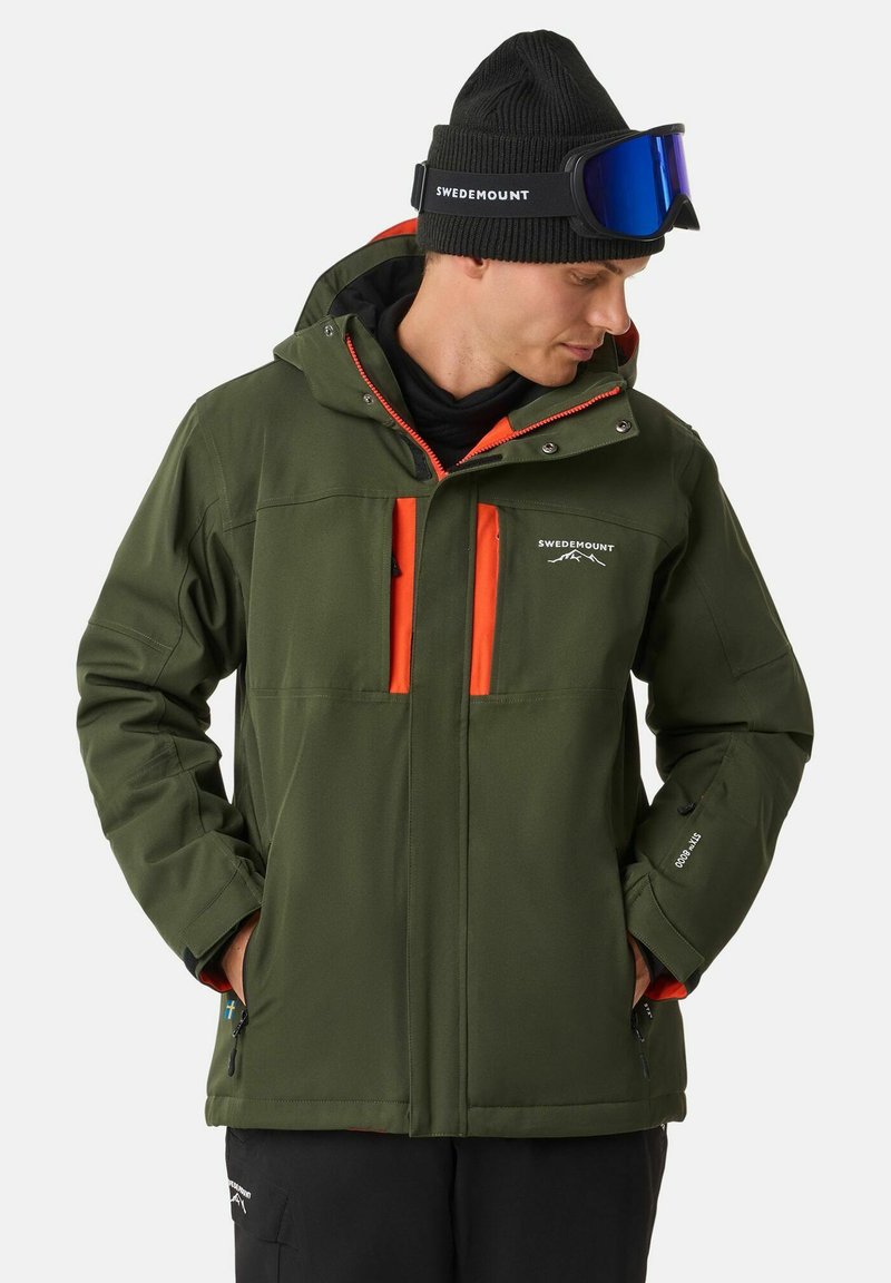Grüne Outdoor-Jacke mit orangefarbenen Akzenten, Reißverschluss-Taschen und Kapuze; kombiniert mit einer schwarzen Mütze und blauen Schutzbrillen. Der Stoff wirkt strukturiert.