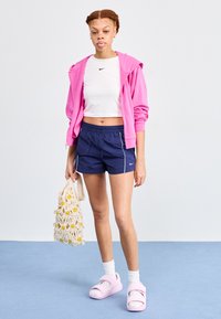 Šviesiai rožinė sportinė striukė, balta trumpa palaidinė, tamsiai mėlyni šortai su baltais kraštais ir rožiniai šlepetės. Laiko nerti maišelį su geltonomis gėlės detalėmis.