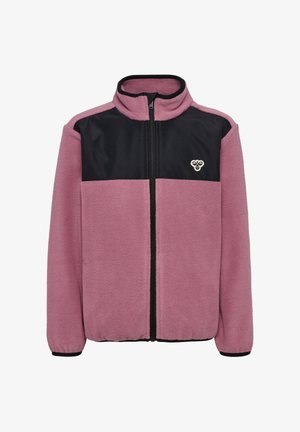 Młoda dziewczyna z zaplecionymi włosami, ubrana w fioletowo-czarną kurtkę na zamek, ciemnoniebieskie dżinsy i zielone trampki, stoi z rękami w kieszeniach.