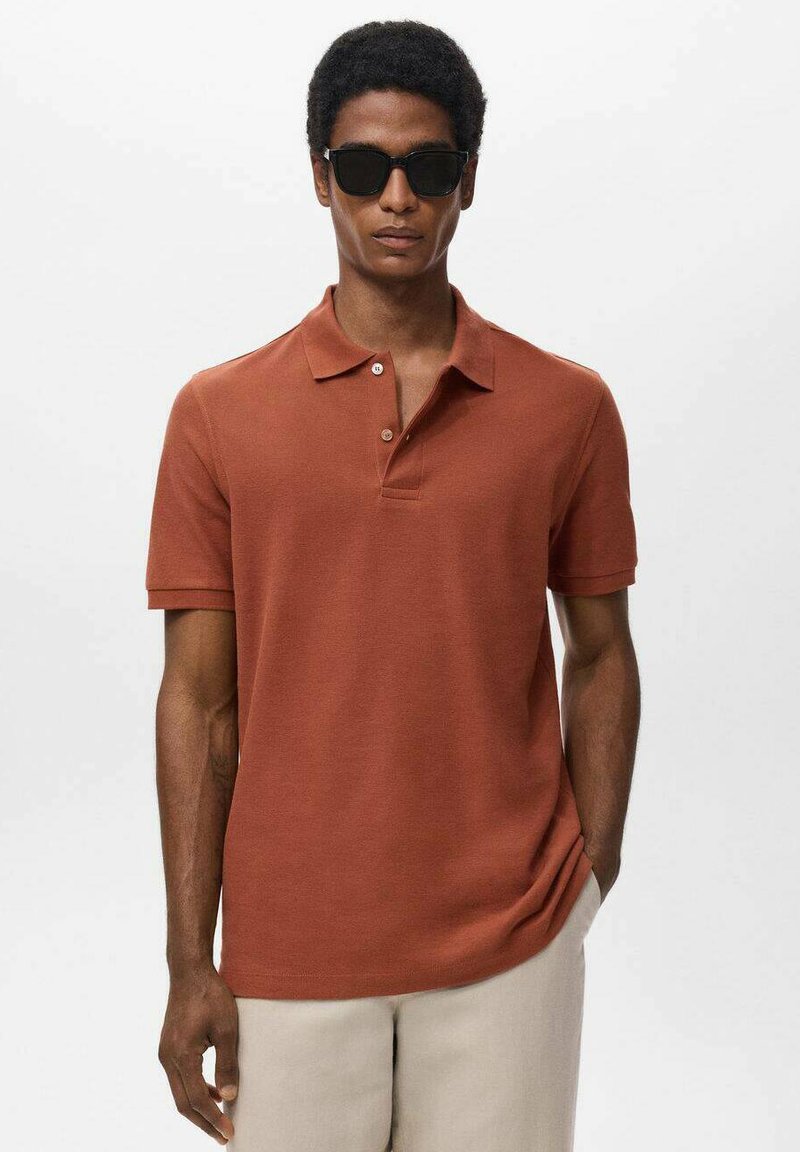 Braunes Polohemd mit einer Knopfleiste mit zwei Knöpfen und kurzen Ärmeln, kombiniert mit hellen Hosen. Modell trägt schwarze Sonnenbrille.