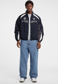 Mørkeblå vindjakke med hvide detaljer og logo, med lynlås foran, høj krave og elastiske cuffs, parret med lyseblå løse jeans.