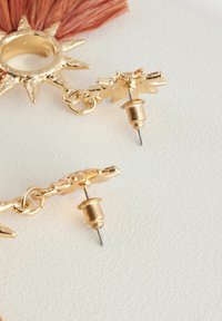 Des boucles d'oreilles dorées au design en éclat de soleil présentent une finition texturée et une attache à clou. Elles incluent un accent en gland rose pour plus de détails.