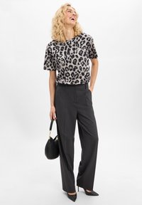 Vrouw in een zwart en grijsgemarmerde luipaardprint shirt, donkergrijze wijde broek, zwarte hakken, die een zwarte handtas vasthoudt, glimlachend en naar boven kijkt.