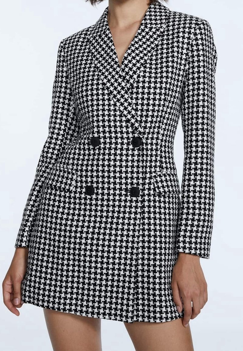 Femme portant une robe blazer croisée à motif pied-de-poule noir et blanc, manches longues et boutons noirs.