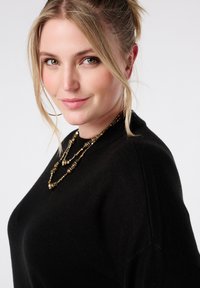 Zwarte trui met een ronde hals, met een zachte textuur. Versierd met een gelaagde gouden kralenketting die licht weerkaatst op de stof.