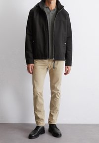 Czarna lekka kurtka z kapturem, pod spodem szara zapinana na zamek bluza, beżowe spodnie oraz czarne sznurowane buty, stojące na neutralnym tle.