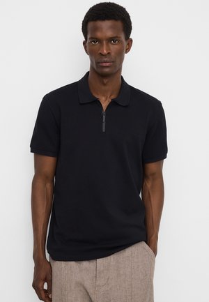 Man draagt een zwart poloshirt met korte mouwen en een rits aan de voorkant en beige broek, staand tegen een effen witte achtergrond.