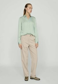 Lys grønn satengbluse med krage, beige skreddersydde bukser og sko med slangeskinnmønster, stående på en nøytral bakgrunn.