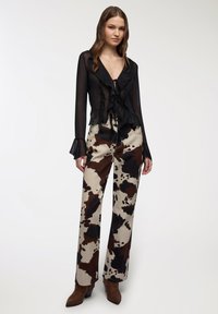 Blusa nera trasparente con dettagli arricciati; pantaloni a gamba larga beige con stampa a macchie di mucca marrone e nera; stivaletti marroni a punta.