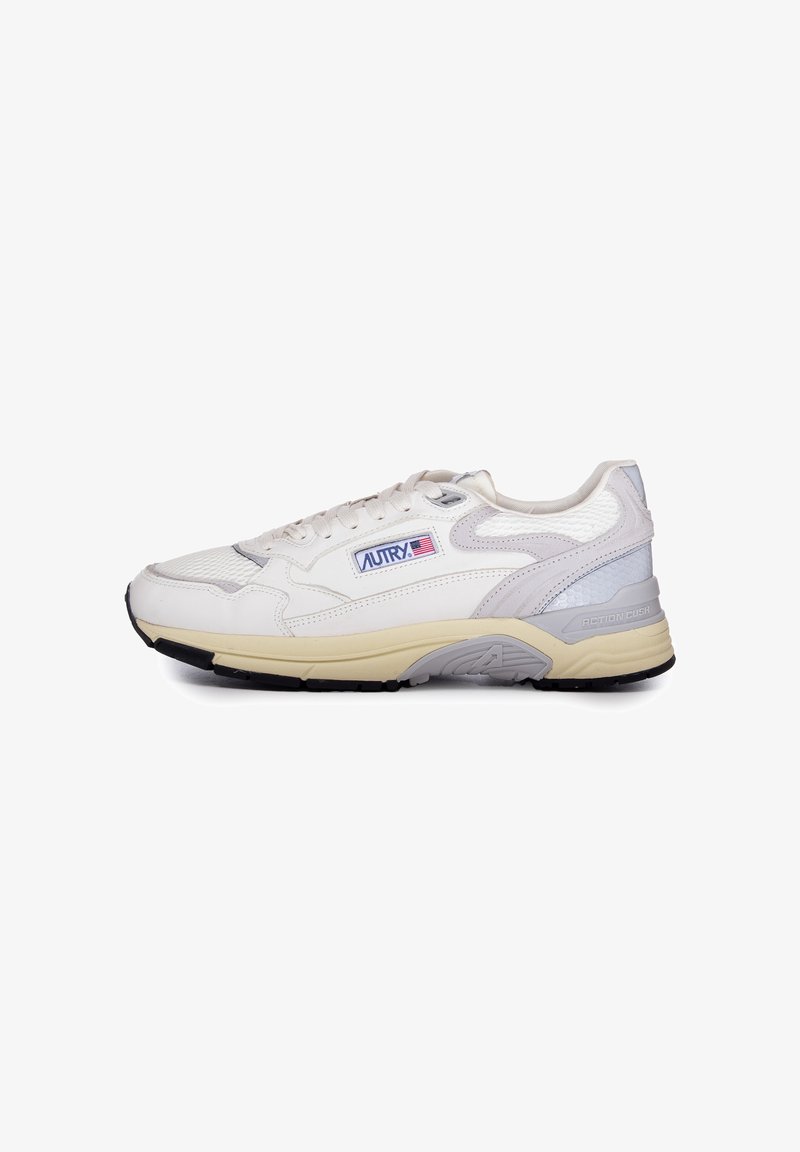 Sneaker bianca realizzata in materiali in rete e pelle. Presenta una suola intermedia beige, dettagli grigi texturizzati e un logo blu e rosso sul lato.