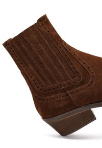 Brązowy zamszowy botek za kostkę z malowniczym designem, poziomym przeszyciem, perforowanymi detalami oraz niskim, warstwowym obcasem.