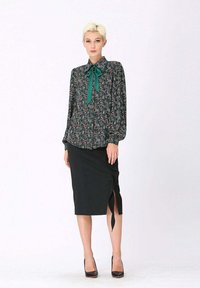 Blouse florale à manches longues avec un nœud vert, associée à une jupe crayon noire avec une fente latérale. Mannequin portant des talons noirs.