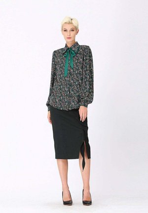 Blouse florale à manches longues avec un nœud vert, associée à une jupe crayon noire avec une fente latérale. Mannequin portant des talons noirs.