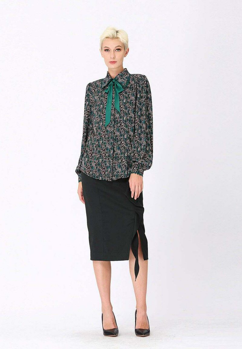 Blouse florale à manches longues avec un nœud vert, associée à une jupe crayon noire avec une fente latérale. Mannequin portant des talons noirs.