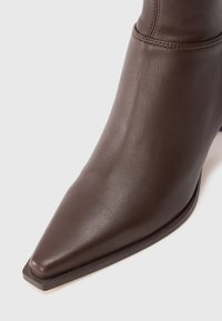 Botine din piele maro închis, cu un design ascuțit și vârful pătrat, având cusături vizibile aproape de marginea superioară.