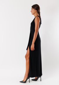 Vestito lungo nero senza maniche con uno spacco laterale, cade morbidamente, caratterizzato da un corpetto aderente. Indossato con tacchi a spillo neri.