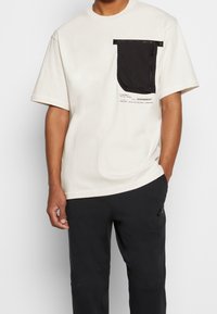 Krémové tričko s krátkým rukávem a černou kapsou se zipem a potiskem. Nosené s černými kalhotami.