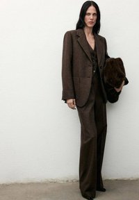 Giacca di abito in lana marrone con rever, abbinata a pantaloni coordinati e un gilet nero con bottoni, mentre tiene un cappotto scuro in pelliccia.
