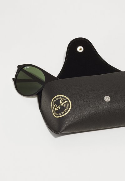 Черни слънчеви очила Ray-Ban, частично видими в черна кожена кутия, с златно лого и сребристо закопчаване. Зелените лещи.