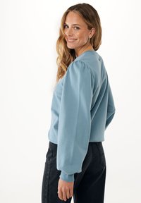 Modrý svetr s balónovými rukávy a žebrovanými manžetami, kombinovaný s černými džíny se zvýšeným pasem a knoflíkovým detailem na boku.