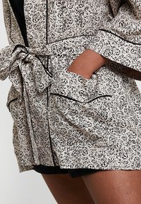 Ropa impresa con un fondo beige y un patrón de hojas negras. Presenta mangas anchas, un cinturón de lazo y ribetes negros en los bordes.