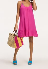 Femme portant une robe sans manches rose vif, tenant un sac de plage tissé avec une serviette rayée rose et jaune, et portant des talons ouverts bleus.