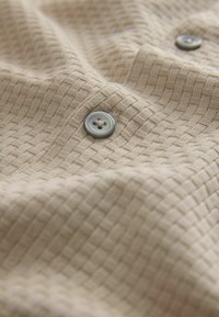 Tissu beige à motif tissé avec deux boutons gris à quatre trous cousus, montrant la texture et de légers plis dans le matériau.