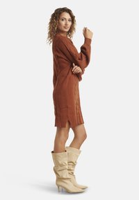 Vestito maglione lavorato a maglia color ruggine con motivi a trecce, maniche lunghe, spacchi laterali, abbinato a stivali beige morbidi al ginocchio.