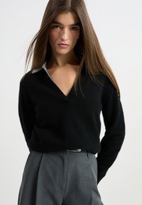 Schwarzer Strickpullover mit V-Ausschnitt und hellgrauem Kragen, kombiniert mit grauen, taillierten Hosen mit gefaltetem Vorderteil.