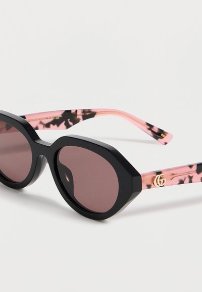 Lunettes de soleil ovales noires avec verres foncés et branches rose écaille avec un logo double-G doré près des charnières.