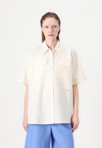 Camicetta corta a maniche corte color crema con due tasche frontali, cuciture a vista e chiusura con bottoni. Abbinata a pantaloni a gamba larga azzurri.