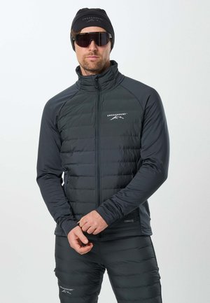 Homme portant un chapeau noir Swedemount, des lunettes de soleil, une veste matelassée et un pantalon, debout devant un fond clair uni.