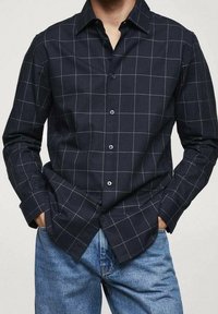 Uomo che indossa una camicia blu navy con motivo a griglia bianca e jeans blu, mani in tasca, su uno sfondo semplice.