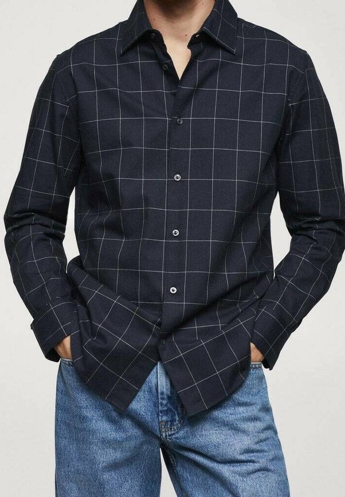 Uomo che indossa una camicia blu navy con motivo a griglia bianca e jeans blu, mani in tasca, su uno sfondo semplice.