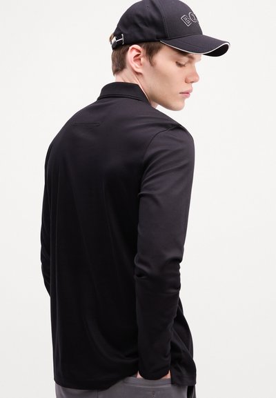 Polo noir à manches longues avec une texture lisse, doté d'un col et d'un subtil détail de couture au dos, associé à une casquette noire.