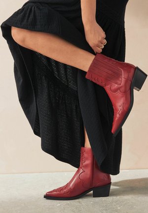 Person hebt einen schwarzen Rock, um kalbhohe rote Lederstiefel mit dekorativer Naht und Blockabsätzen zu zeigen.