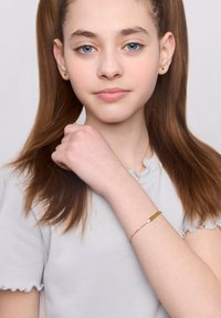 Złota bransoletka z prostokątnym charms'em na delikatnym łańcuszku, noszona na nadgarstku. Modelka ma proste brązowe włosy i nosi jasnoszary top.